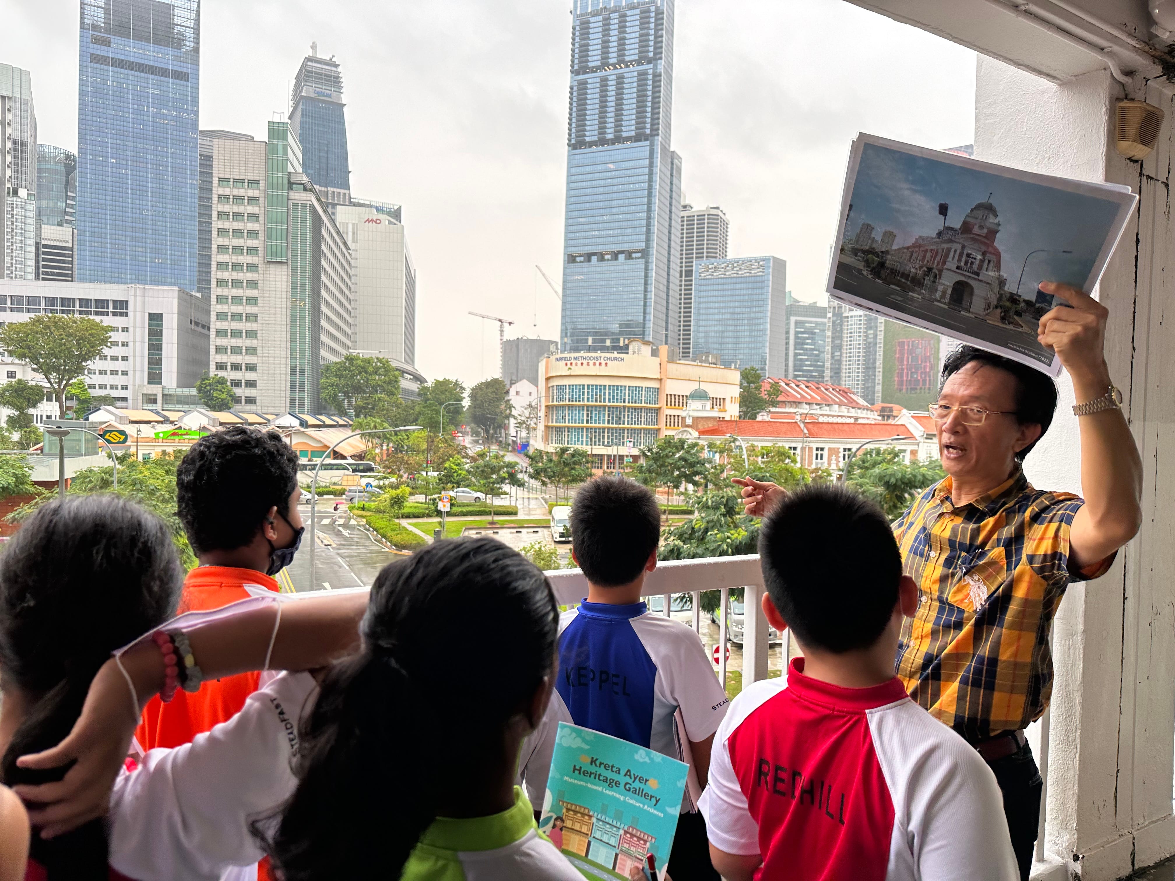 Tour guide holds up a photo while talking to a group on a balcony overlooking city skyscrapers.